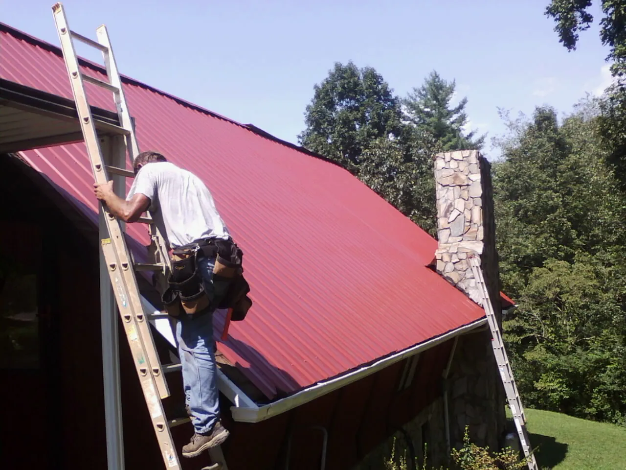 Roofer on a ladder beside a red metal roof and stone chimney — Soffit & Fascia Repair by Roofing Brothers in Berlin Center